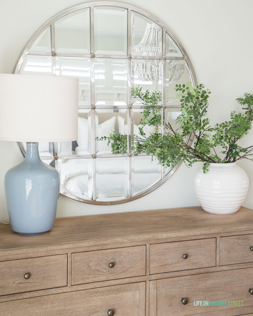 A large silver round mirror with windowpane details hung above a light wood dresser. The dresser is decorated with a blue gray lamp and white ceramic vase filled with artificial greenery stems.