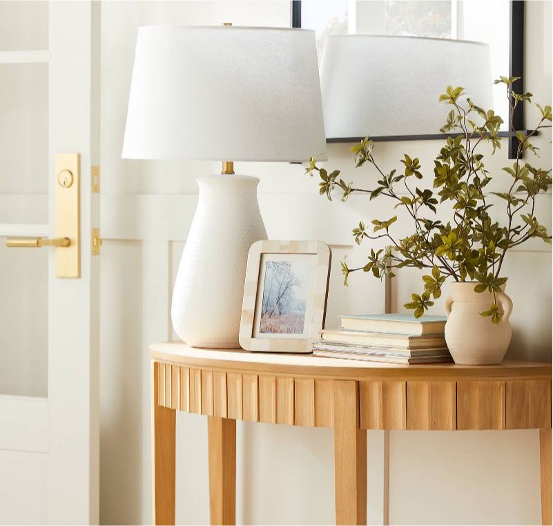 New Releases From Studio McGee including a light wood scalloped demilune console table, a white ceramic lamp, black mirror, picture frame, and faux greenery.