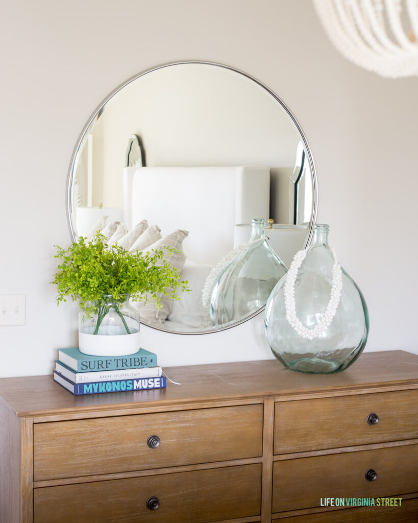 An oversized round mirror above a wood dresser in a neutral guest bedroom.