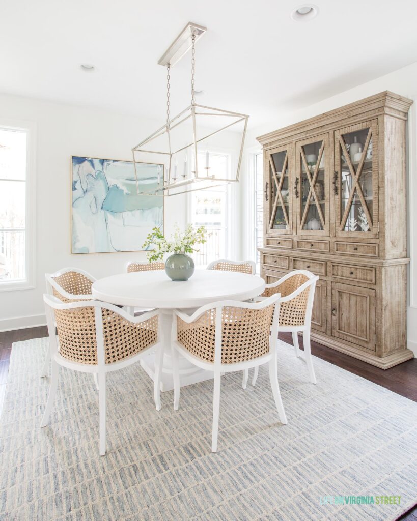 A guide to modern coastal round dining tables. This dining room features the Serena & Lily Terrace Round Dining Table with cane backed chairs, a blue and cream rug, a large wood hutch, a linear chandelier, and coastal abstract art.