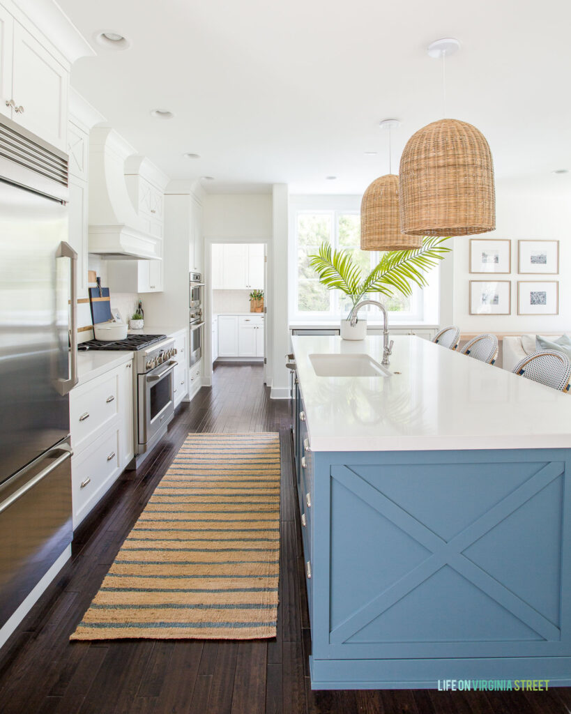 A coastal kitchen with a blue kitchen island, basket pendant lights, white perimeter cabinets, and Caesarstone Calacatta Nuvo quartz for the kitchen countertops.