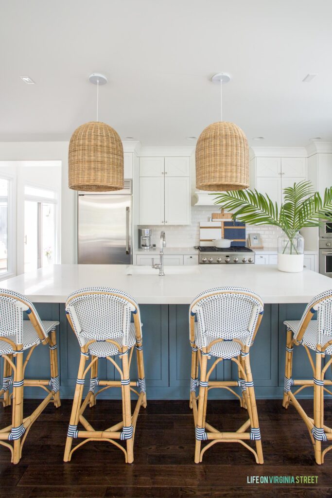 A coastal kitchen with white cabinets and dark blue island with four bistro style counter stools and woven basket pendant lights.