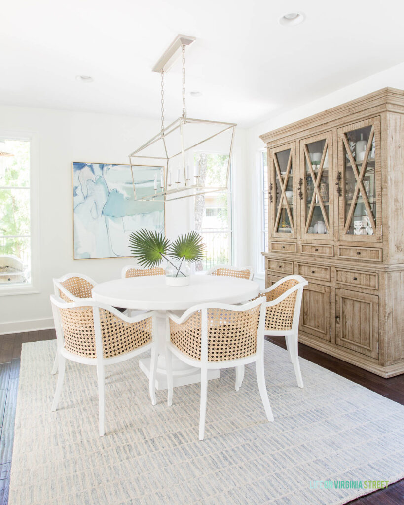 A dining room decorated for summer with a white round concrete dining table, cane dining chairs, a light wood hutch, a blue and cream patterned rug, faux palm fronds, oversized blue abstract art, and a linear chandelier.