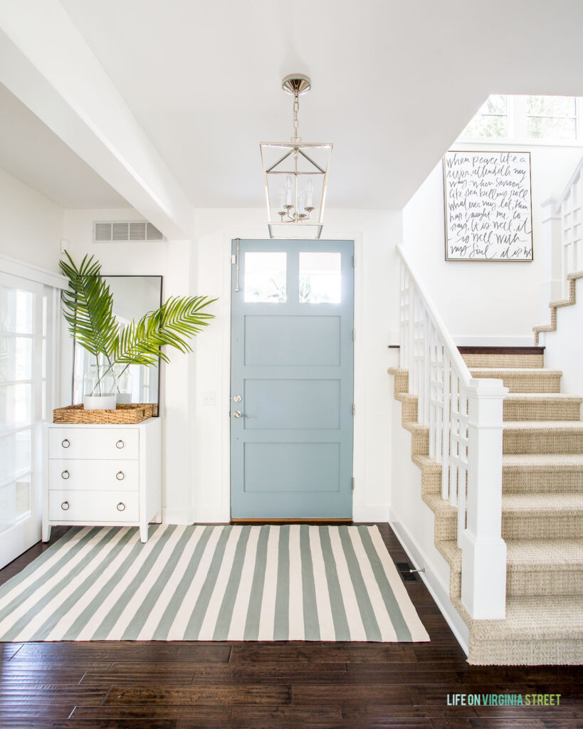 A light and bright entryway with a white raffia cabinet, blue striped rug, palm leaf fronds, a light blue front door, lantern pendant light, and "It Is Well" art hung in a stairwell.