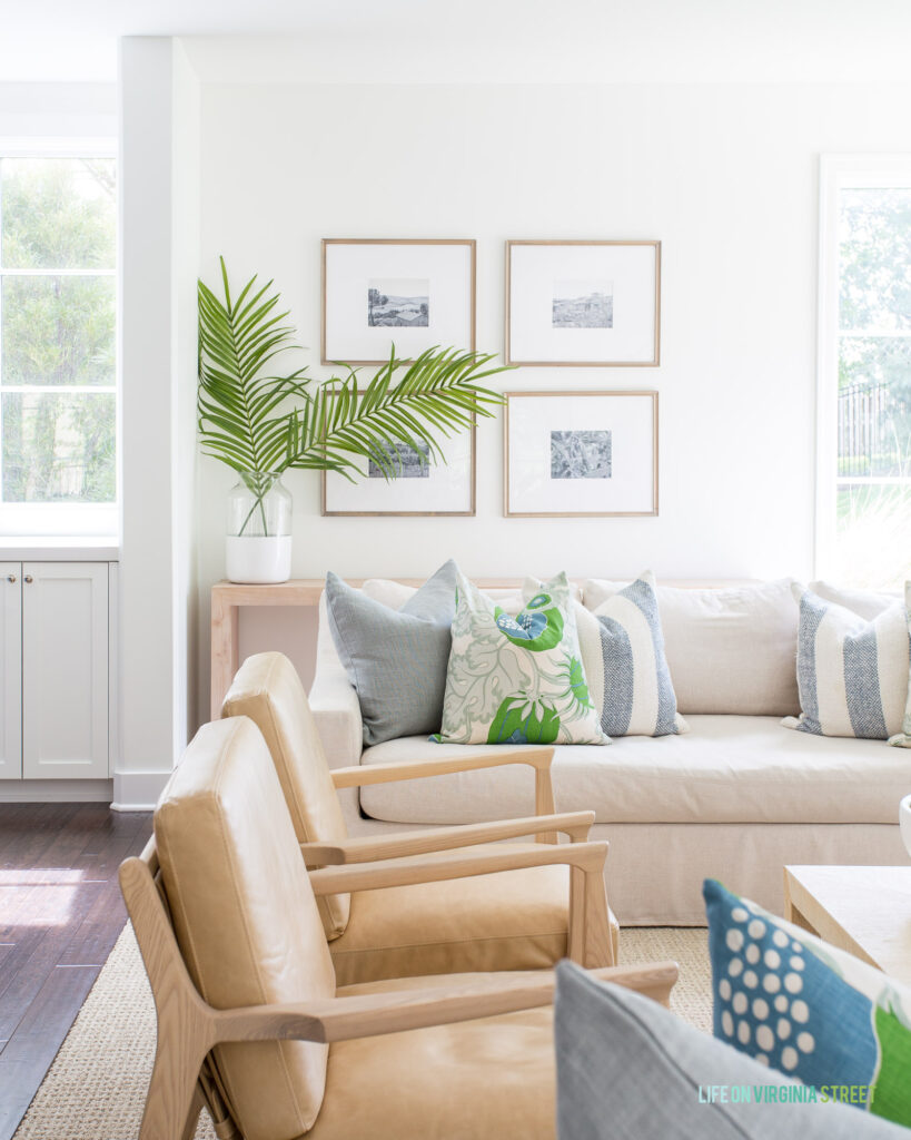A cozy summer living room feature a linen sofa, light leather armchairs, wood gallery wall frames, blue and green throw pillows, palm leaf branches, and natural light from large windows.