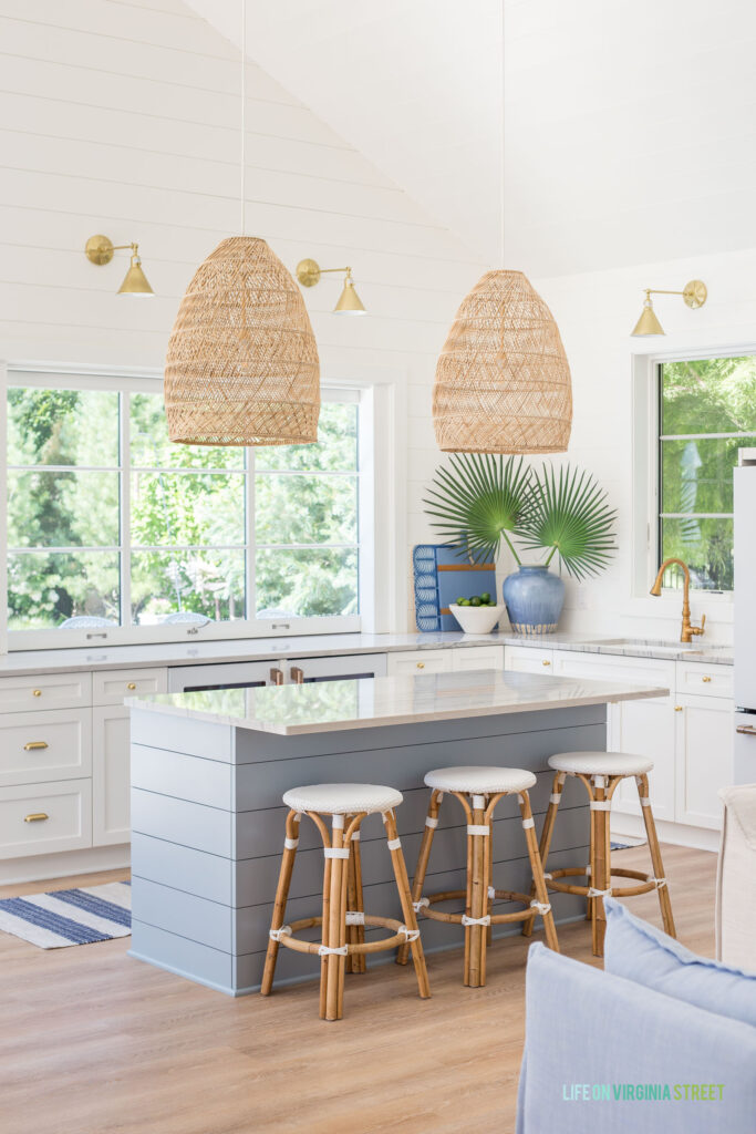 A coastal style pool house kitchen featuring three white and rattan backless counter stools at the island. The island is painted Benjamin Moore Santorini Blue and has woven pendant lights hanging above it.