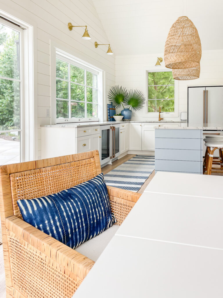 A woven rattan dining chair in a coastal inspired pool house with basket pendant lights, a blue striped rug, brass sconces and an island painted Benjamin Moore Santorini Blue.