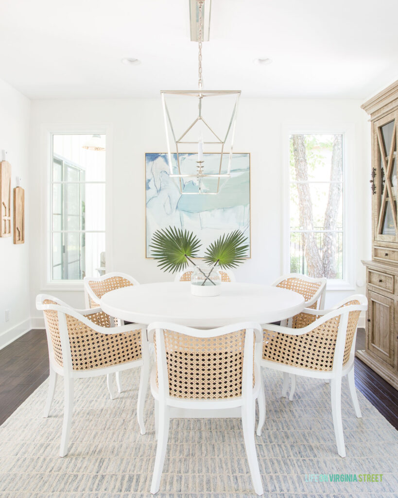 Modern coastal cane dining chairs in a dining room with a blue patterned rug, linear chandelier, abstract art, faux fan palms, a white dining table, and large wood hutch.
