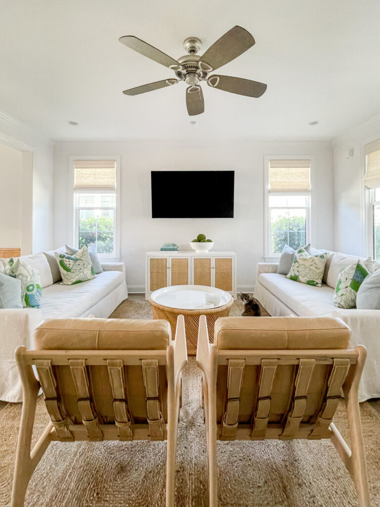 A Florida coastal cottage living room with linen sofas, light leather chairs, a jute rug, rattan cabinet, and round coffee table. Decorated with blue and green accents.