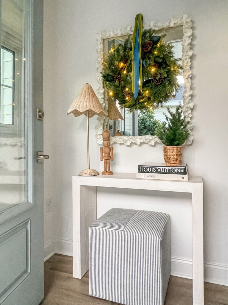 A small entryway decorated for Christmas with a light wood console table, striped ottoman cube, rattan table lamp, wreath with blue and green velvet ribbon, a wood nutcracker, a coral mirror, and a mini tree in a seagrass basket.