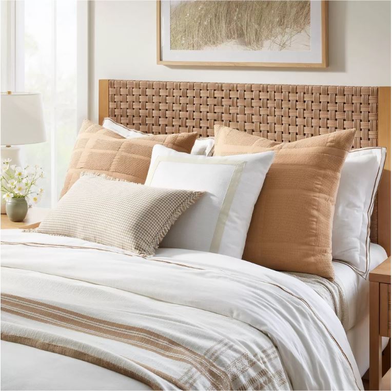 Bedding from the Studio McGee 2023 spring line includes a woven headboard, layers of white and copper colored bedding, a check throw pillow and cosmos in a floral arrangement.