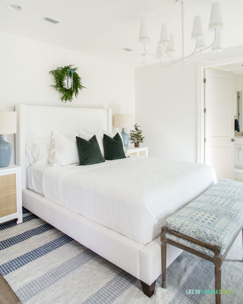 A 2022 Christmas home tour featuring a neutral bedroom with a white headboard, blue striped rug, wreath hung over the bed, dark green velvet pillows, a white chandelier, rattan nightstands and blue gray ceramic lamps.