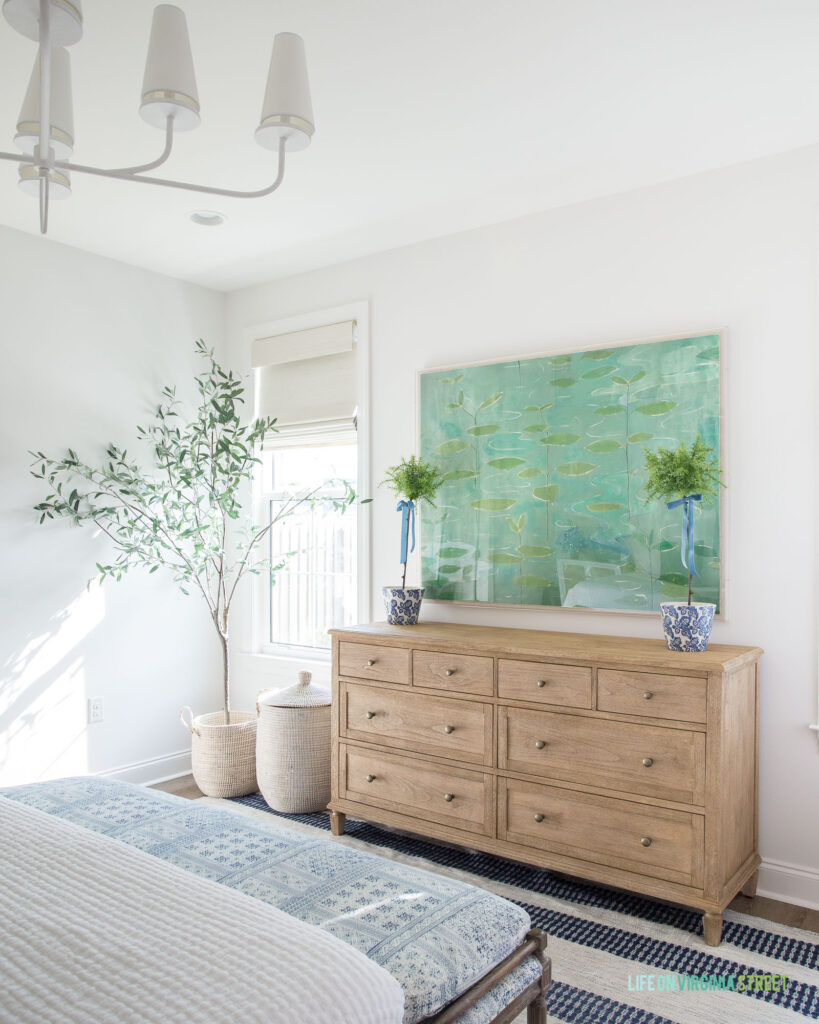 A coastal bedroom with a light wood dresser, blue and green lily pad art, topiaries with light blue velvet ribbons, a white chandelier, an artificial olive tree, blue striped rug, and batik style bench.