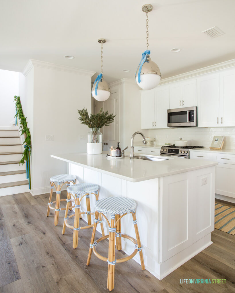 A small white kitchen decorated for Christmas with silver pendant lights with light blue velvet bows, woven backless counter stools, a vase filled with evergreen stems, and garland on the staircase.