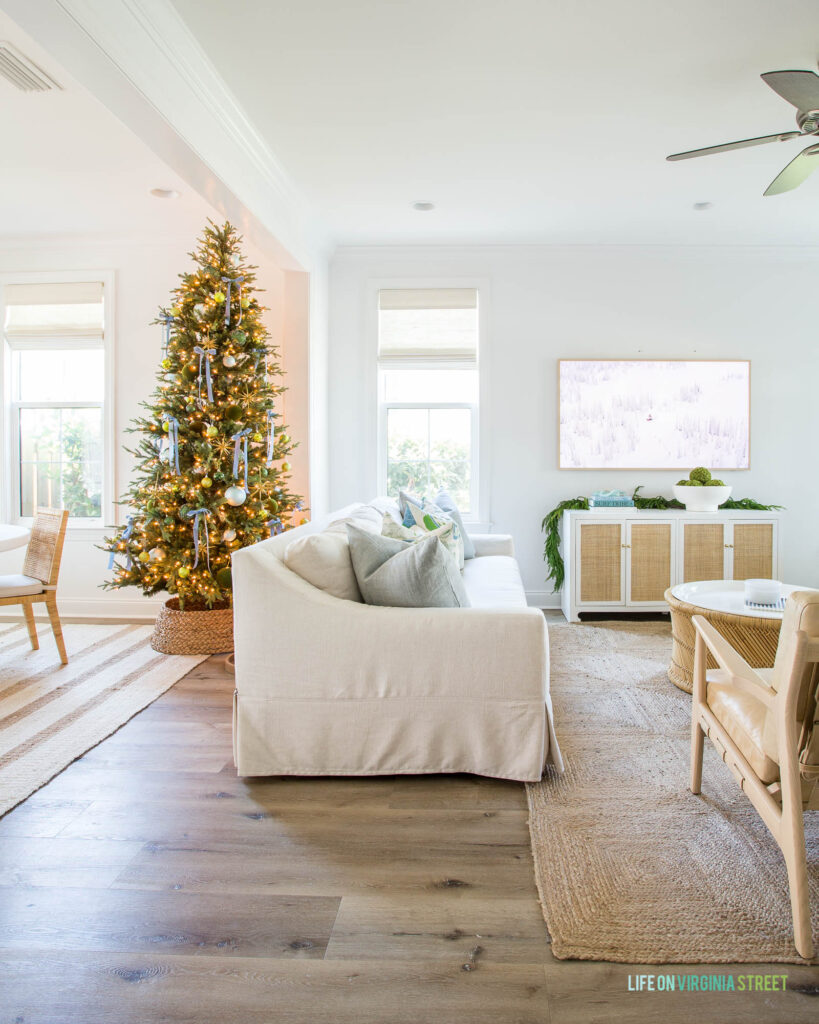 A coastal living room and dining room with a tall Christmas tree, linen sofa, jute rug, light blue velvet ribbon, and rattan media console, Frame TV, and a seagrass tree collar.