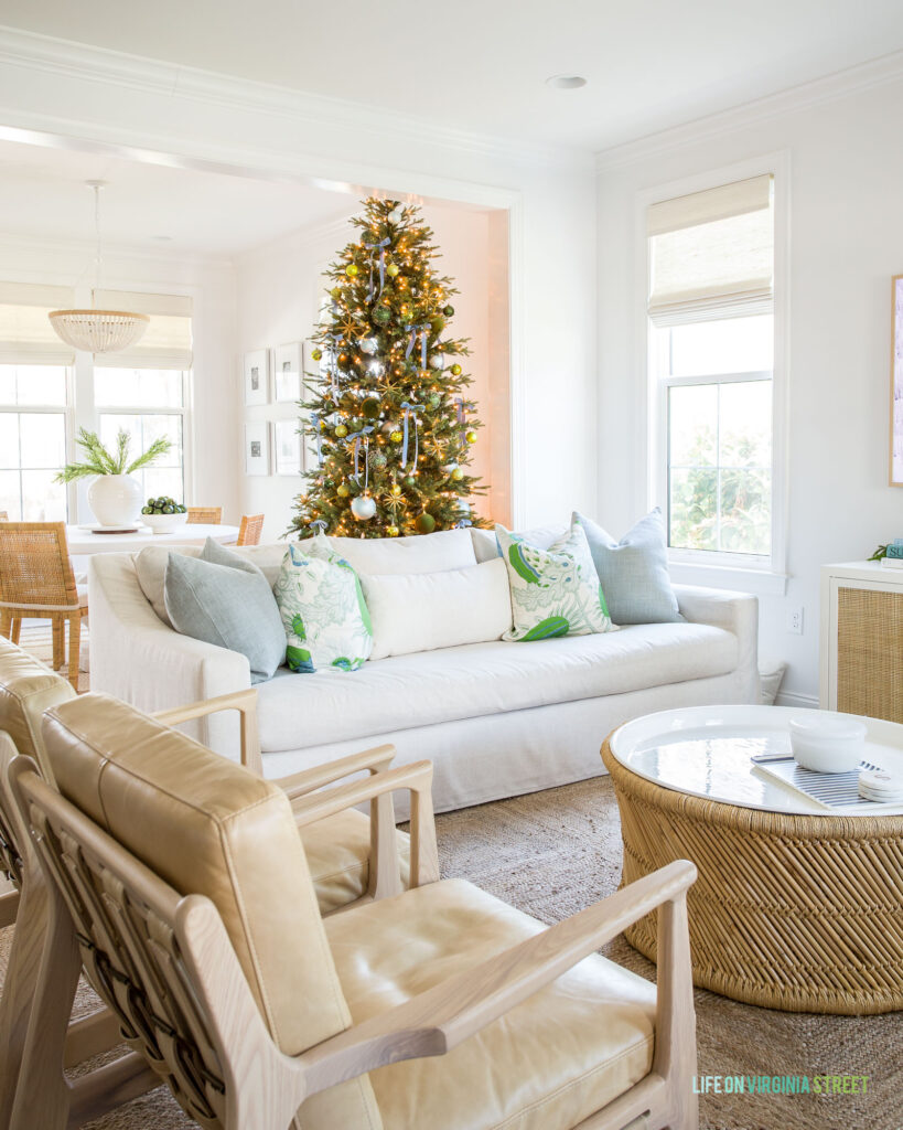 A coastal living room decorated with blue and green throw pillows, light leather armchairs, a round bamboo coffee table, a 9' artificial Christmas tree and a jute rug.