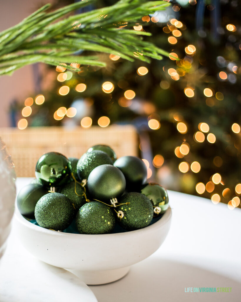 A simple white footed bowl filled with leftover green ornaments. You can see the lights from the Christmas tree in the background.