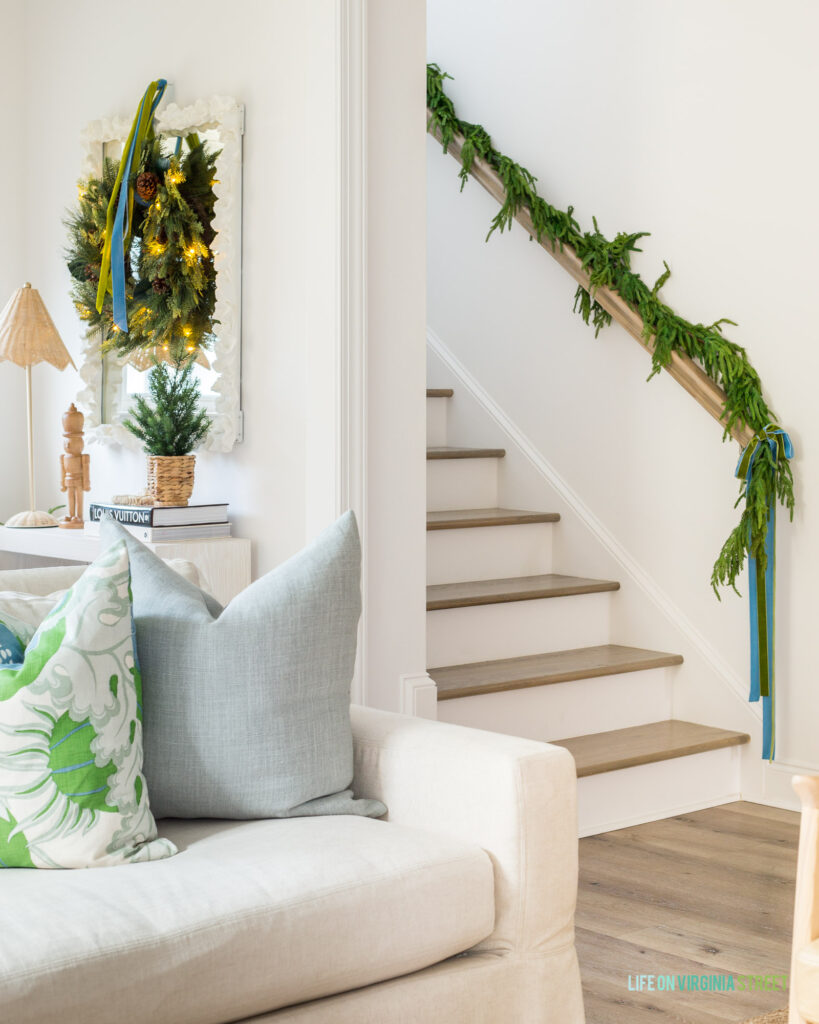 A Christmas home with a linen sofa, blue and green throw pillows, garland on the staircase, a small console table, a wreath with Christmas lights and a rattan table lamp.