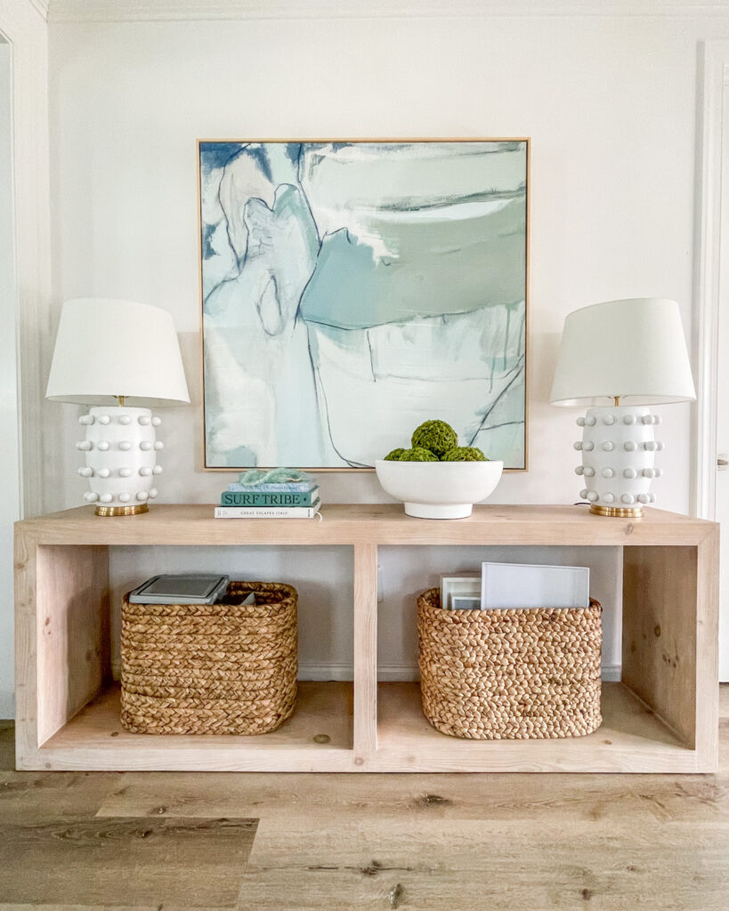Abstract coastal art hung above a light wood console table. Styled with white dot lamps, seagrass baskets, and coastal coffee table books.