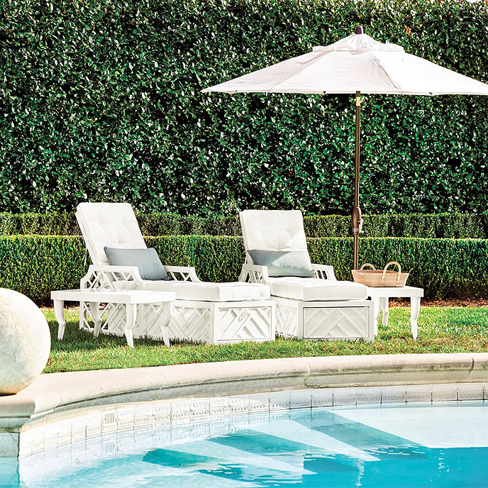 White Bermuda style chippendale chaise lounge chairs styled with a white umbrella around a light blue pool.