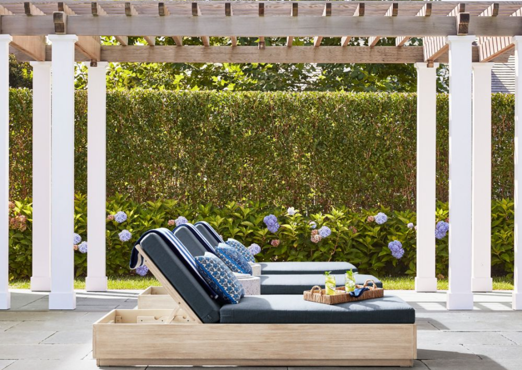 Eucalyptus wood outdoor lounge chairs with navy blue cushions under a large pergola near a hedge with blue hydrangeas.