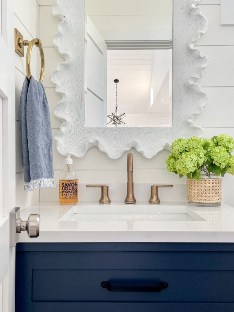 A white wavy coastal bathroom mirror in a bathroom with white shiplap walls, a navy blue vanity, gold hardware, and simple floral arrangement on the countertop.