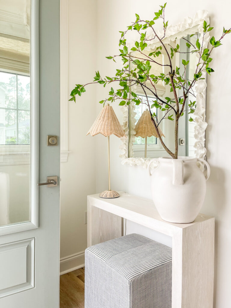 A large white ceramic vase filled with a faux tree on a narrow console table in a coastal entryway.