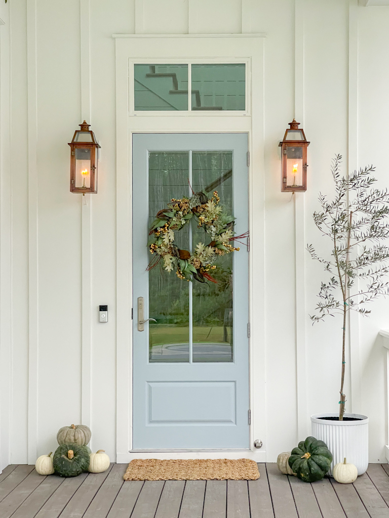 A fall front porch with green pumpkins, an olive tree in a white fluted pot, and copper lanterns. The board and batten house is painted Sherwin Williams Greek Villa and the front door is painted Sherwin Williams Niebla Azul.