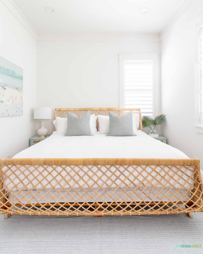 A cozy Florida carriage house bedroom with the rattan Serena & Lily Avalon bed, a light blue herringbone rug, beach art, light blue and cane nightstands, faux fan palm leaves, and a white bedside table lamp.
