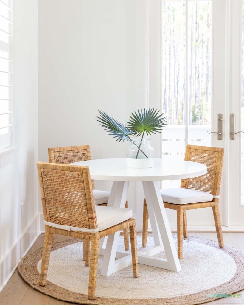 A Florida carriage house dining space with a white round dining table, rattan dining chairs, a round jute rug, and fan palms on the table.
