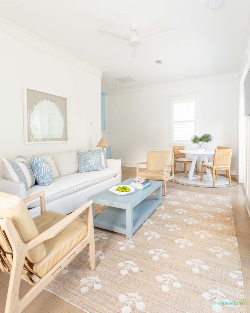 A Florida carriage house living room with the walls and trim painted Sherwin Williams Snowbound. The decor features fan palm art, a light slipcovered sofa, light leather chairs, a block print rug, light blue coffee table, and a white ceiling fan.