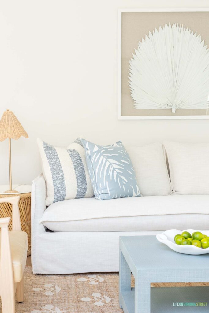 Our Florida carriage house living room decorated in coastal blues and neutrals. Includes a slipcovered sofa, fan palm art, a light blue coffee table, palm print pillows, a rattan scalloped lamp and block print rug.