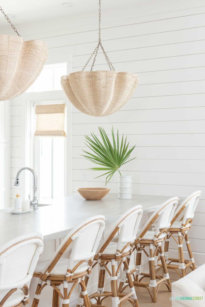 A coastal kitchen with white shiplap walls, Serena & Lily white Riviera swivel counter stools, Palacek Isla chandeliers over the kitchen peninsula, a striped Carrara marble vase filled with saw palms, and woven Roman shades on the window from blinds.com.