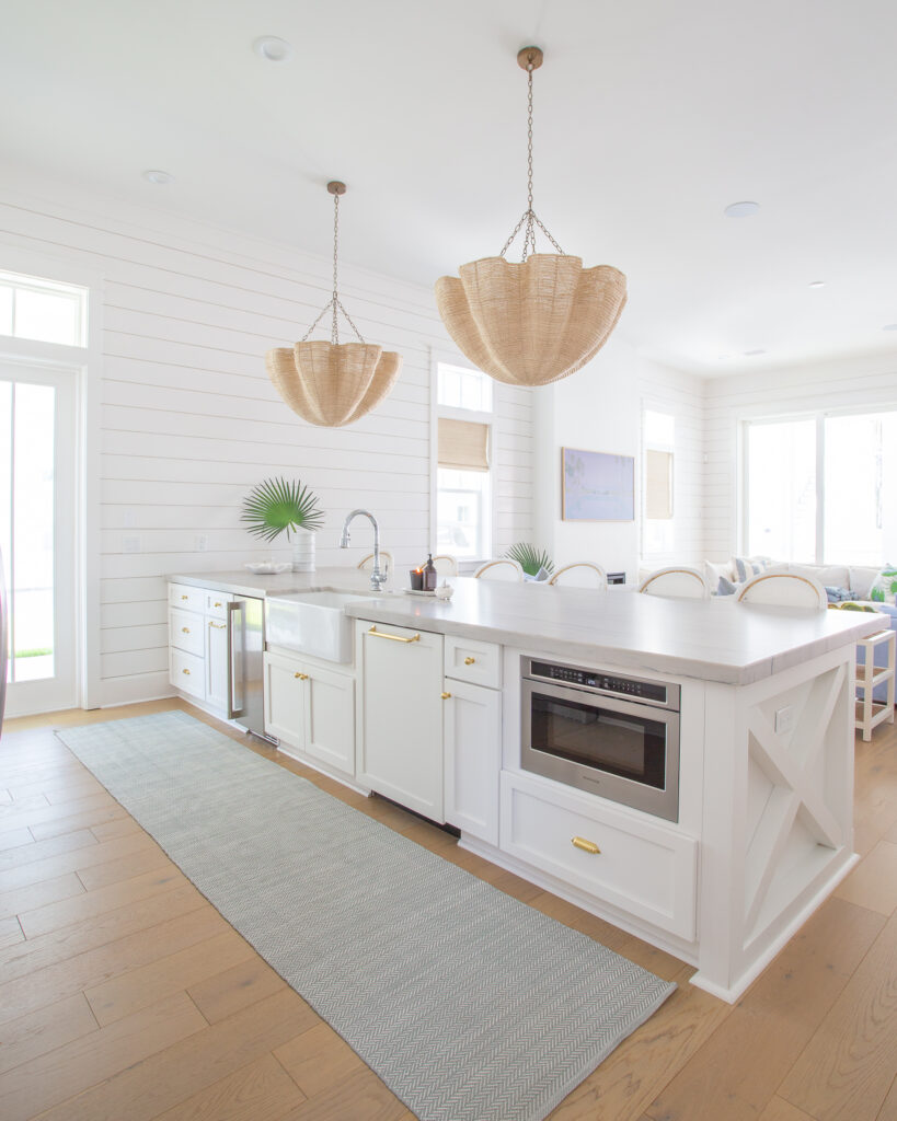 A coastal kitchen with warm white cabinets, white shipalp walls, woven scalloped Palacek pendant lights, white swivel counter stools, and light blue accents.