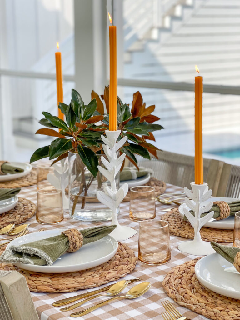 Fluted orange candlestick tapers on a simple Thanksgiving tablescape with buffalo check tablecloth, amber drinking glasses, gold flatware, olive green napkins, water hyacinth chargers and napkin rings, and freshly clipped magnolia stems as a centerpiece.