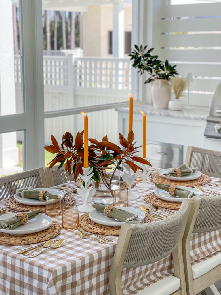 A simple Thanksgiving tablescape idea with a taupe buffalo check tablecloth, water hyacinth placemats, rope dining chairs, gold flatware, amber drinking glasses, magnolia stems and orange tapered candles.