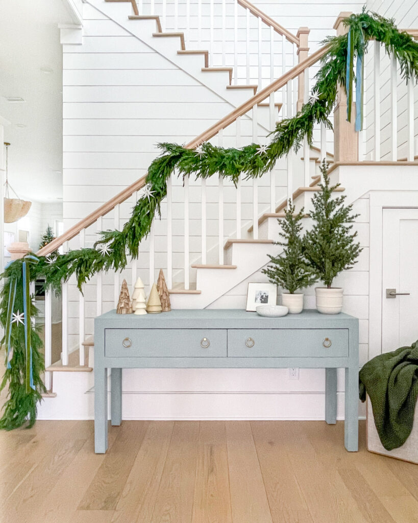 Target Christmas decor finds styled in a bright entryway with white shiplap wall, light blue console table and light oak floors. Accessories include garland down the staircase, faux mini Christmas trees, a cozy throw blanket, and velvet ribbon.