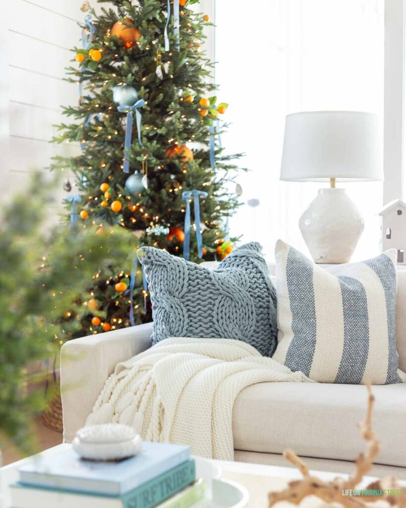 Citrus Christmas decor in a neutral living room with light blue velvet ribbons, faux orange stems, cozy throw pillows and blankets, striped blue pillow and a 9' Christmas tree.