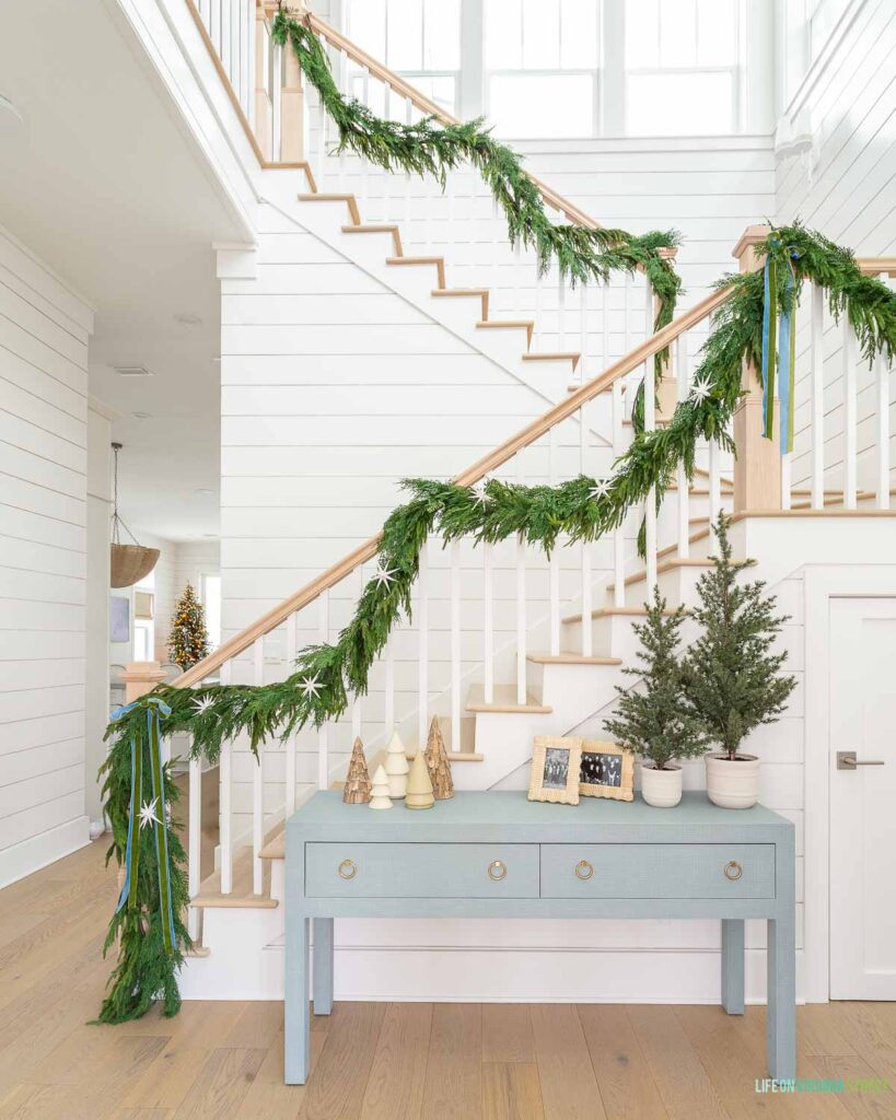 A coastal Christmas home entryway with white oak hardwood floors, white shiplap walls, a blue linen console table, garland on the staircase, and neutral and blue accents.