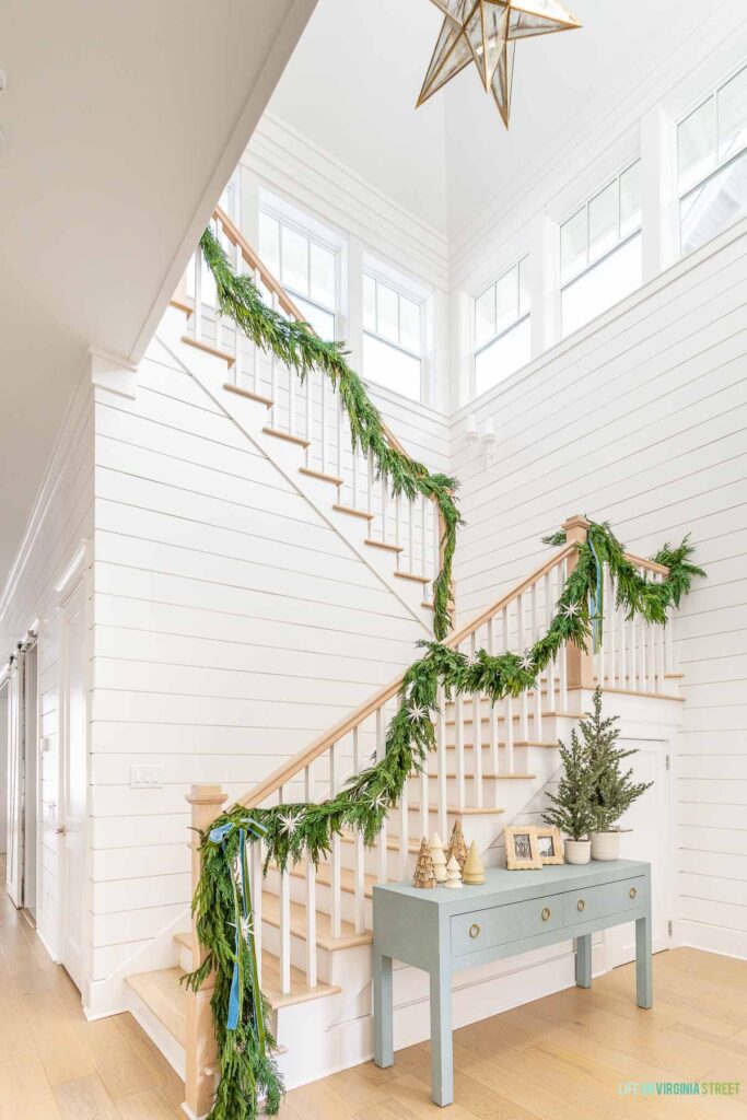 Evergreen garland tied to a staircase with white oak floors and white shiplap walls. A large gold chandelier hangs in the space.