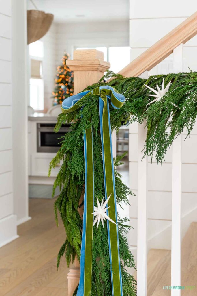 Blue and green velvet ribbon tied on Christmas garland on a staircase with white oak handrail and white spindles.