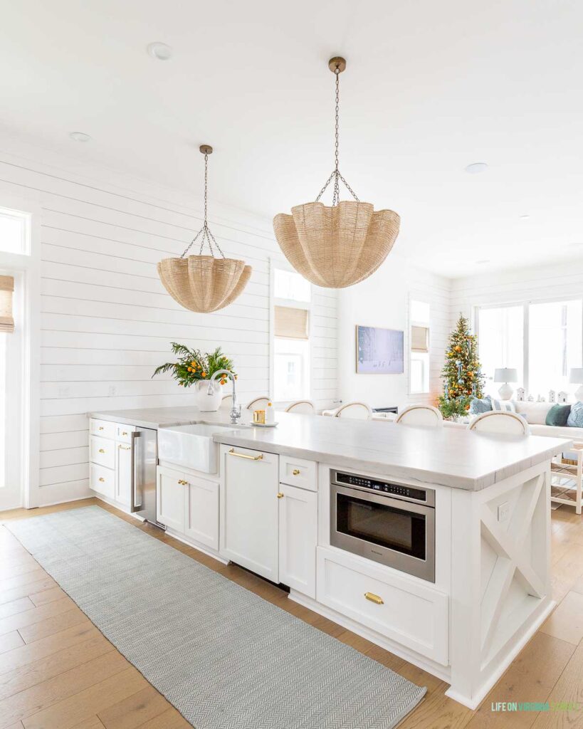 A coastal home kitchen with oversized Palecek scalloped chandeliers, a herringbone rug, white cabinets, gold hardware, and a Christmas tree in the background.