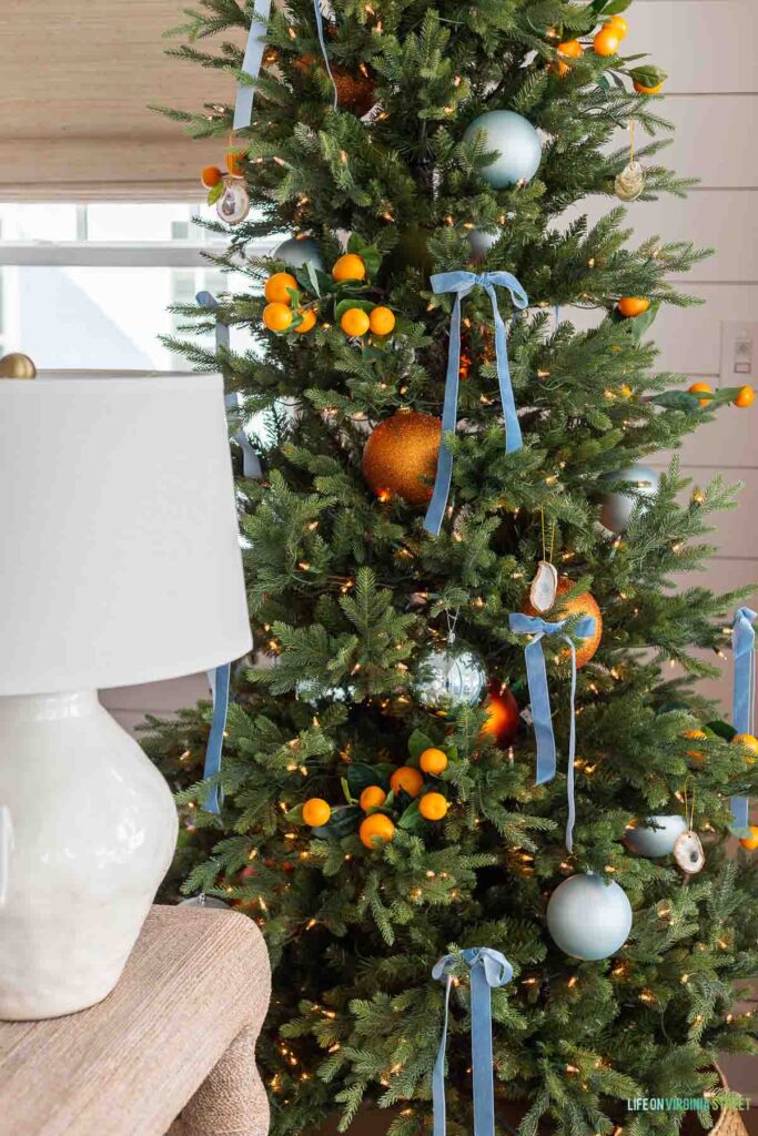 A coastal Christmas tree decorated with orange stems, light blue orb ornaments, large orange ornaments, oyster shell ornaments, and light blue velvet ribbon bows!