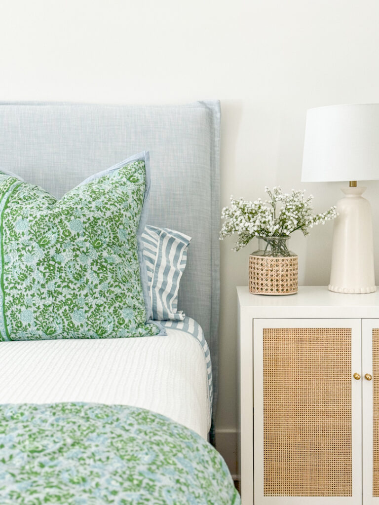 This primary bedroom includes a blue linen bed from Serena & Lily, white waffle knit blanket, a blue and green quilt, blue and white striped sheets, rattan nightstands, faux baby's breath in a cane wrapped vase, and a jute rug.