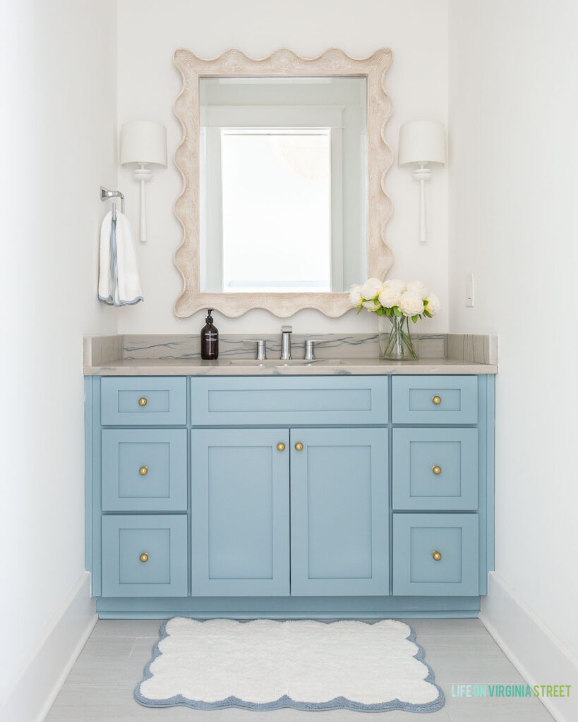 A coastal bathroom decorated with light blue cabinets, gold cabinet knobs, a scalloped wood mirror, white sconces, a blue and white scalloped bath mat, and faux peonies for spring.