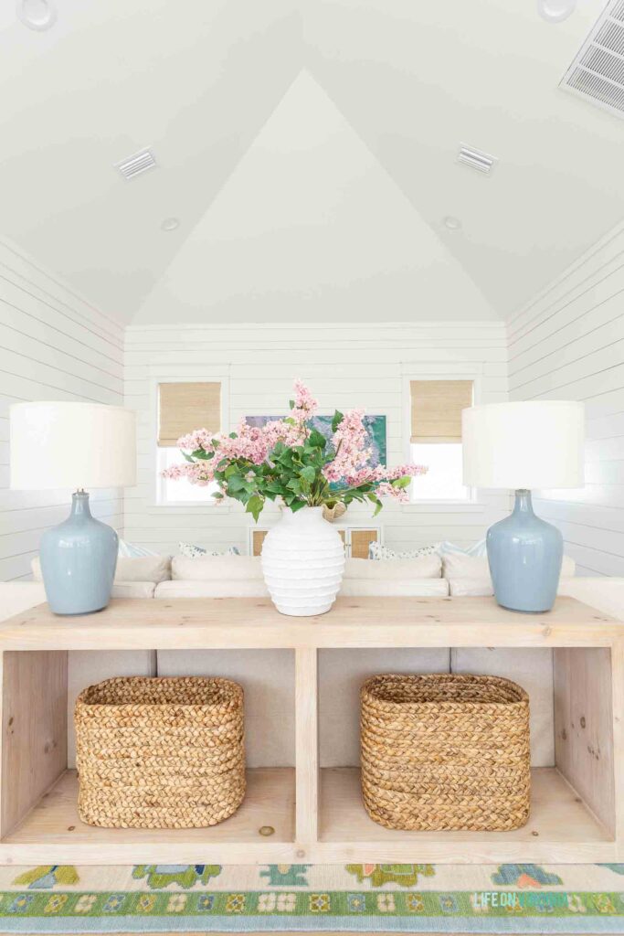 Loft living room decorating for spring! The room includes a vaulted ceiling with white shiplap walls, a long wood console table, blue gray table lamps, a linen sectional, colorful oushak rug, and faux pink lilac stems.