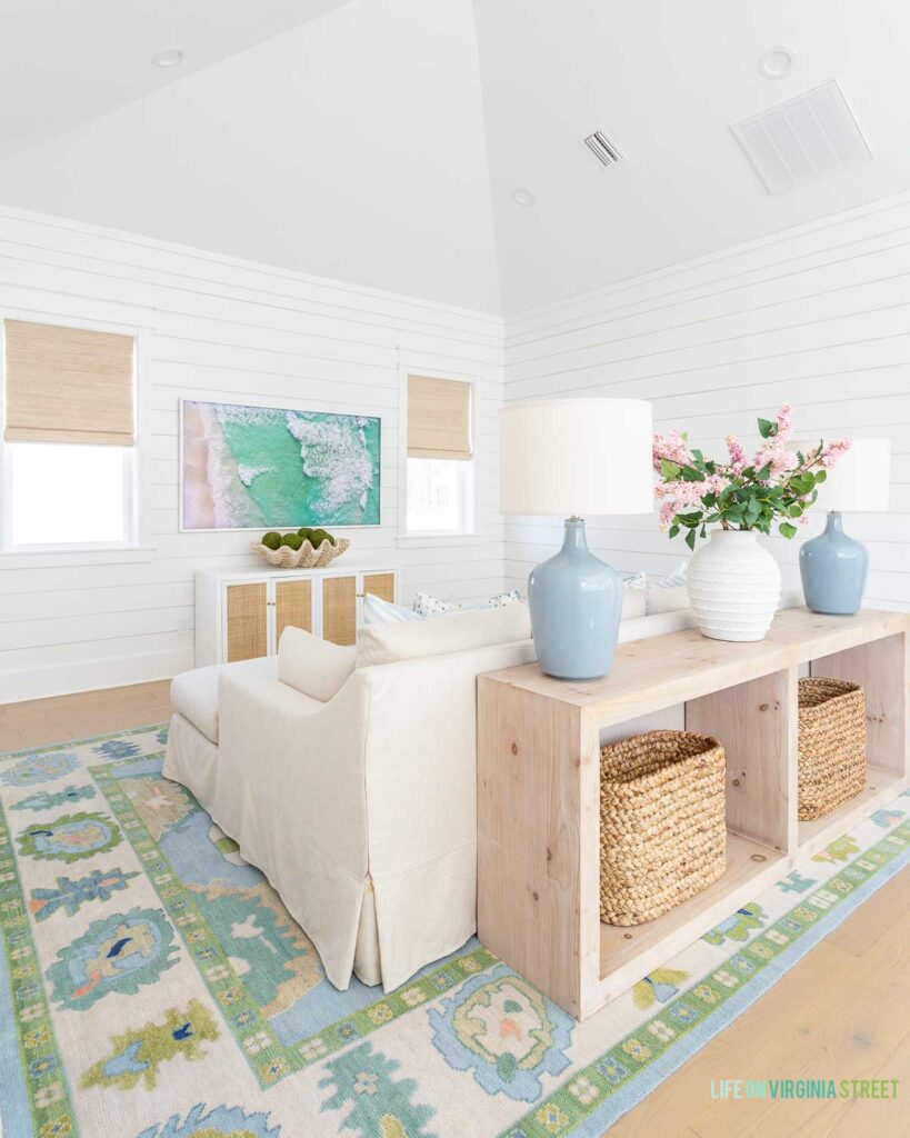 Loft living room decorated for spring! The room includes a vaulted ceiling with white shiplap walls, a long wood console tablewith woven baskets, blue gray table lamps, a linen sectional, coastal art on the Frame TV, cane cabinet, colorful oushak rug, and faux pink lilac stems.