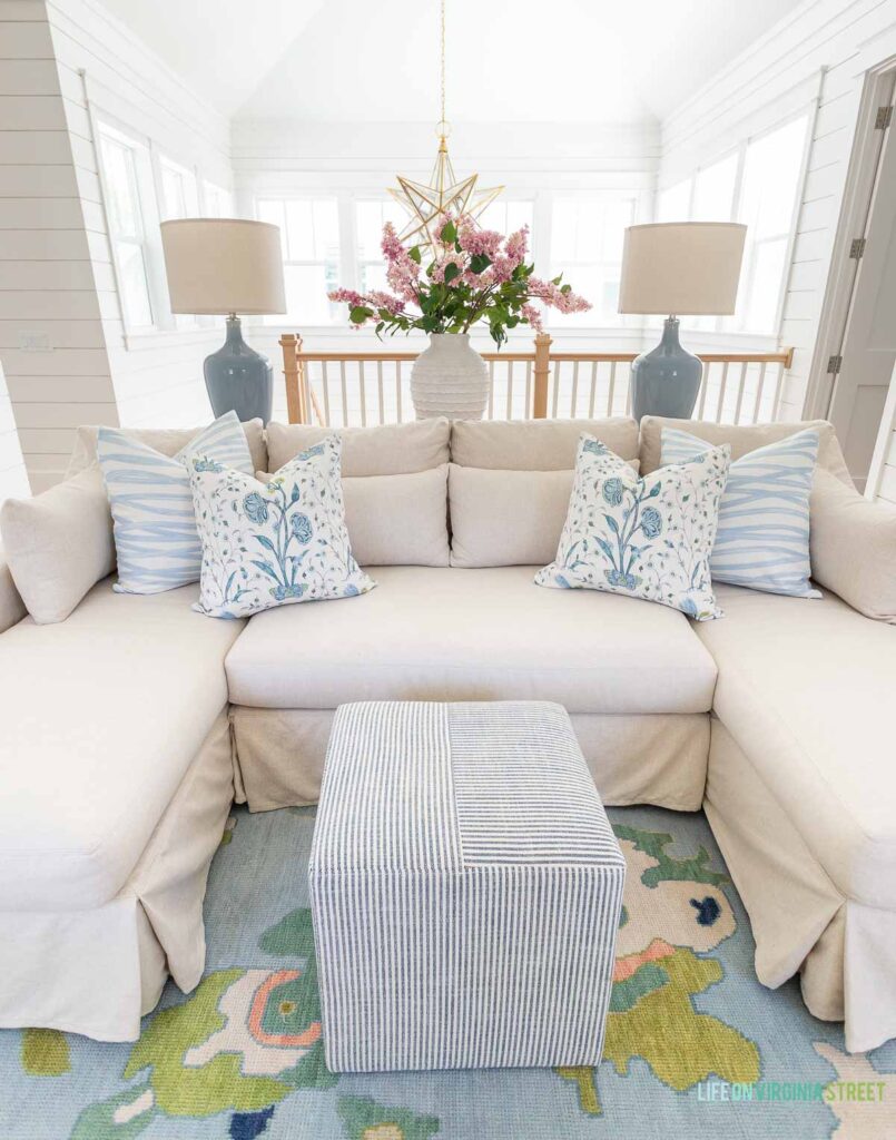 Loft living room decorated for spring! The room includes a vaulted ceiling with white shiplap walls, blue gray table lamps, Moravian star chandelier, a linen sectional, colorful oushak rug, blue brushtroke pillows, floral pillows, a striped ottoman and faux pink lilac stems.