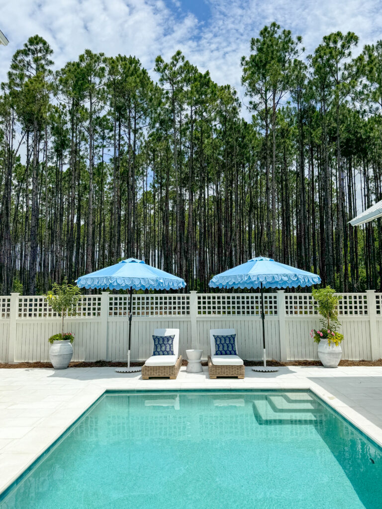 The Life On Virginia Street pool details for her northwest Florida pool. This is a Pebble Tec pool with light pavers, light blue scalloped umbrellas, light woven pool chairs, a white wood fence, and pots filled with citrus topiaries and flowers.