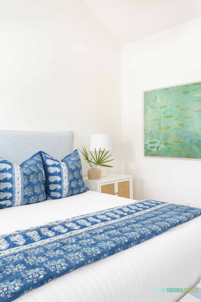 A coastal bedroom with a white jute rug, light blue upholstered bed, vaulted ceiling, blue summer bedroom decor, a white and rattan nightstand, and a cane wrapped vase filled with saw palm fronds.